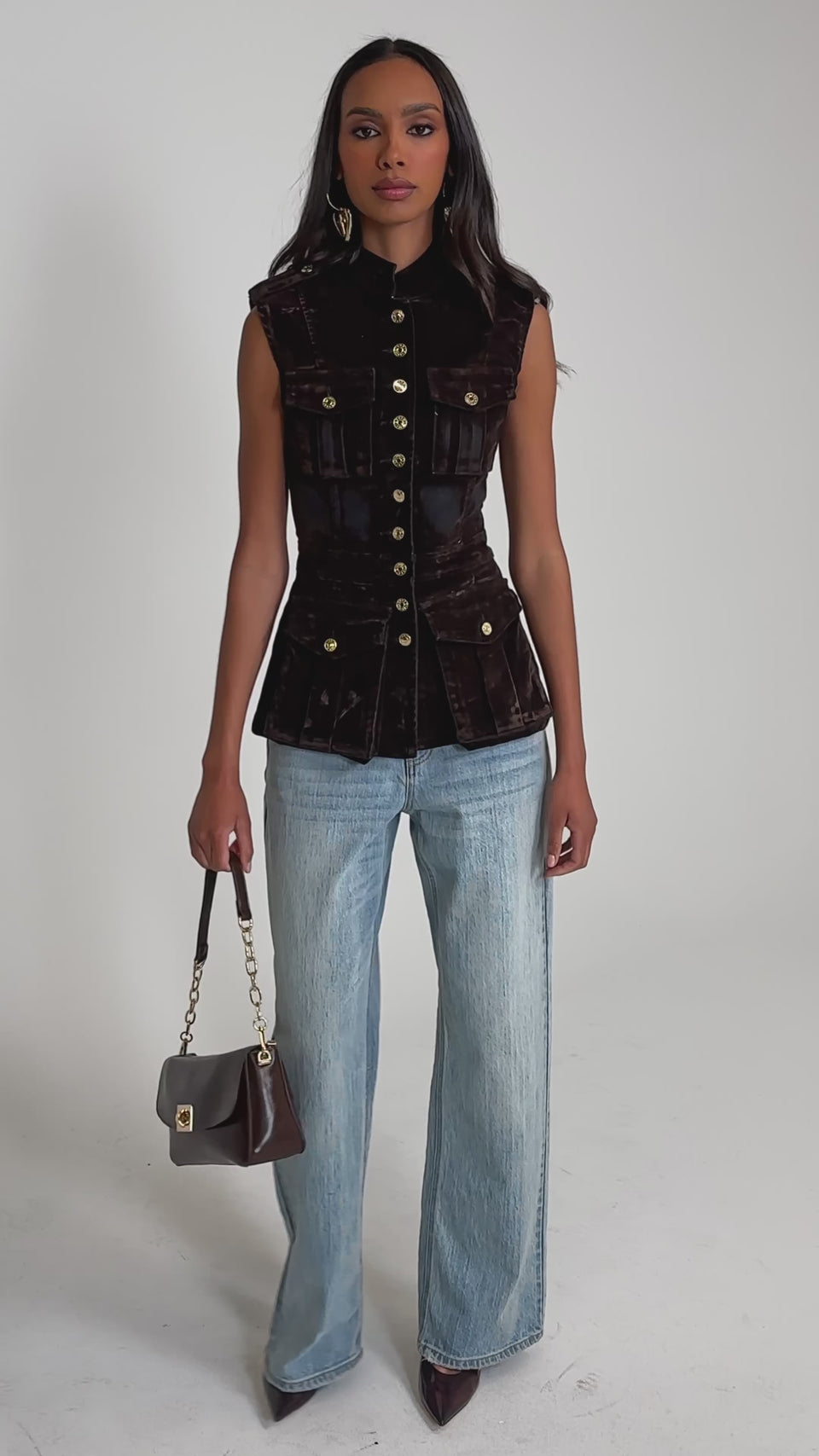 Video of woman modeling the BACK WITH IT VELVET BUTTON DOWN, a fitted brown military-style vest with gold buttons and flap pockets, paired with light blue jeans against a plain light gray background.