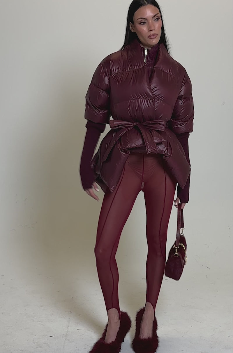 A video of a woman in the RIB SLEEVE PEPLUM PUFFER IN BURGUNDY, paired with sheer burgundy leggings, furry heels, and a matching small bag. A plain background highlights her bold, fashion-forward style.
