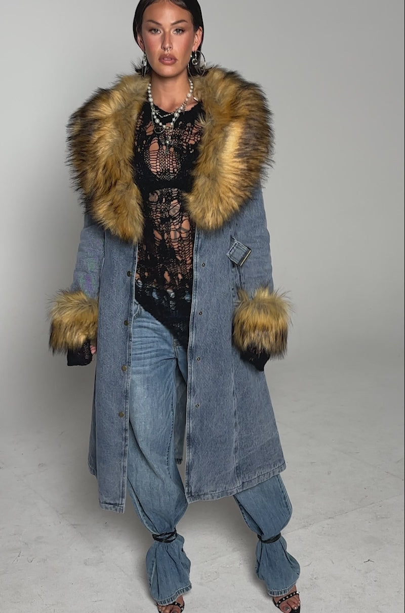 A video of a woman wearing the KAYA FUR TRIM DENIM TRENCH, featuring a faux fur collar and cuffs, over a black lace top and wide-leg jeans. 