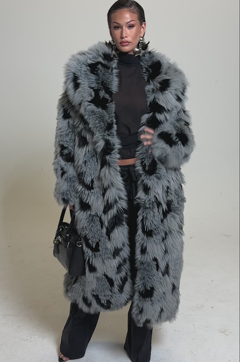 A video of a person wearing the APRES SKI FAUX FUR COAT in a longline black and gray spotted design, paired with black pants and holding a black handbag.