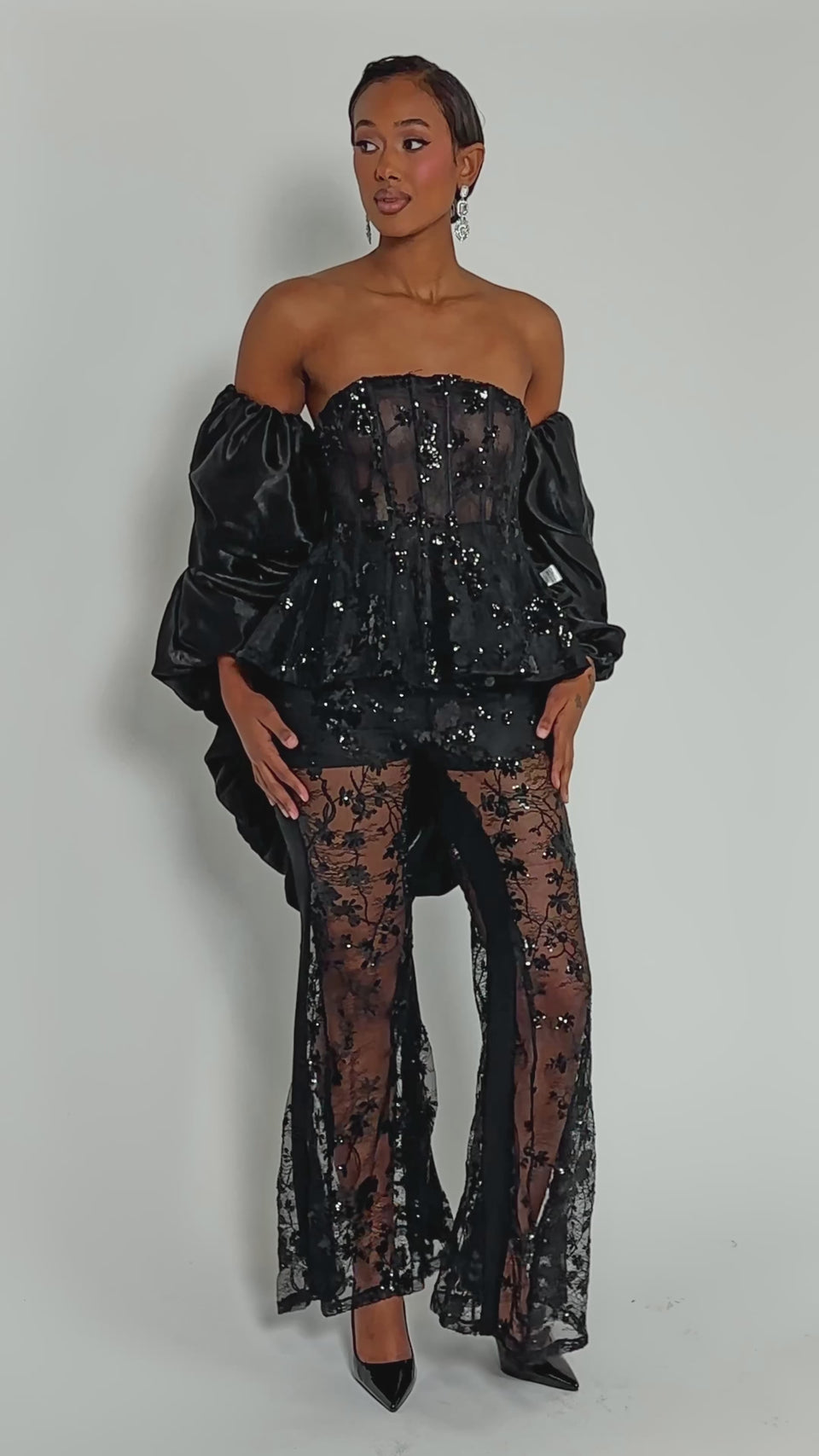 Video of woman modeling the MORE THAN ENOUGH SATIN SHRUG IN BLACK over a strapless black gown with a ruched back and lace details, accessorized with dangling earrings, against a plain background.