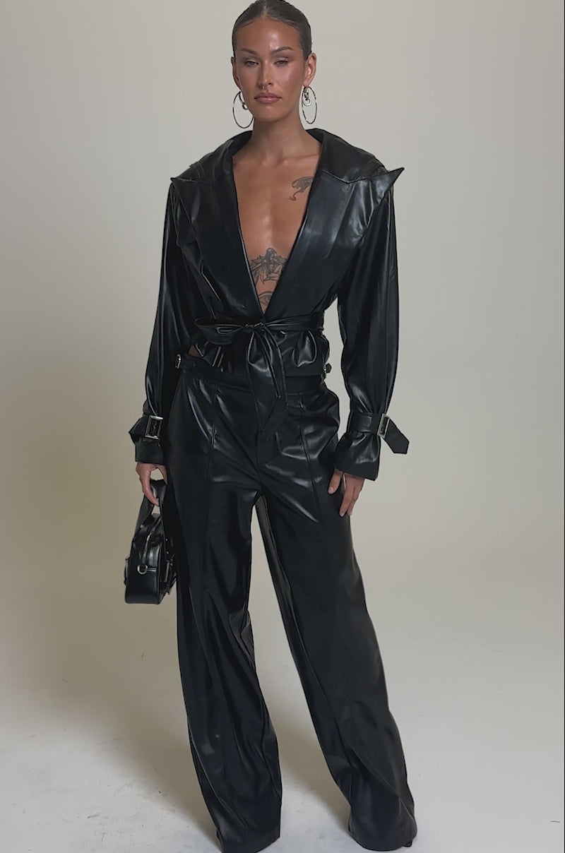 Video of woman modeling the MONTE CARLO HOODED MOTO IN BLACK and matching faux leather pants poses confidently with a small black handbag. Her open jacket shows a chest tattoo, and hoop earrings finish her bold look.