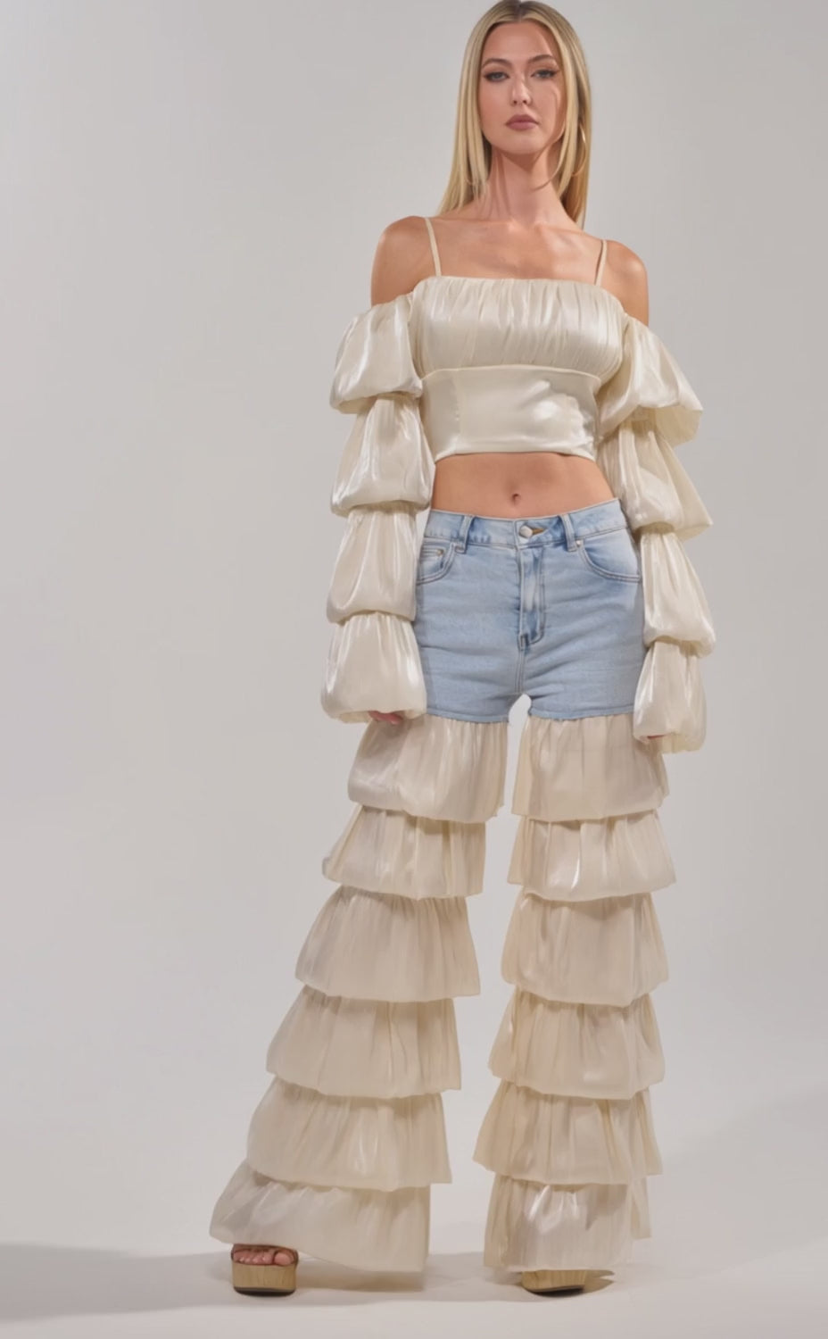 Video of woman modeling the  BOUNCIN AROUND BLOUSE—an ivory off-shoulder crop top with tiered sleeves—styled with light blue denim shorts and a chain belt, standing against a plain background.