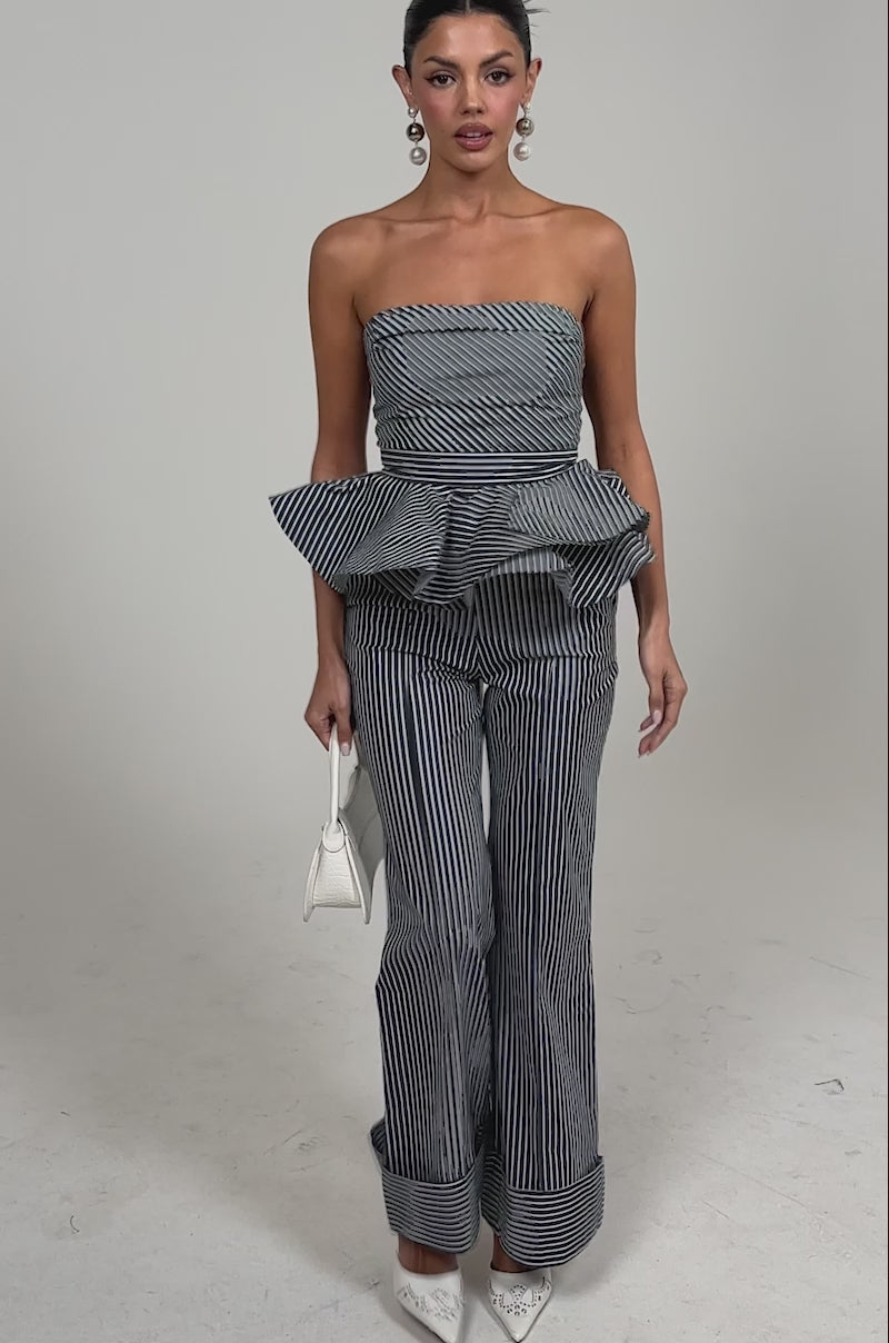 Video of woman modeling the MISS POPULAR POPLIN TROUSER, a pair of navy and white wide-leg pinstripe pants, paired with a matching strapless peplum top. She accessorizes with white heels, a white handbag, statement earrings, and slicked-back hair.