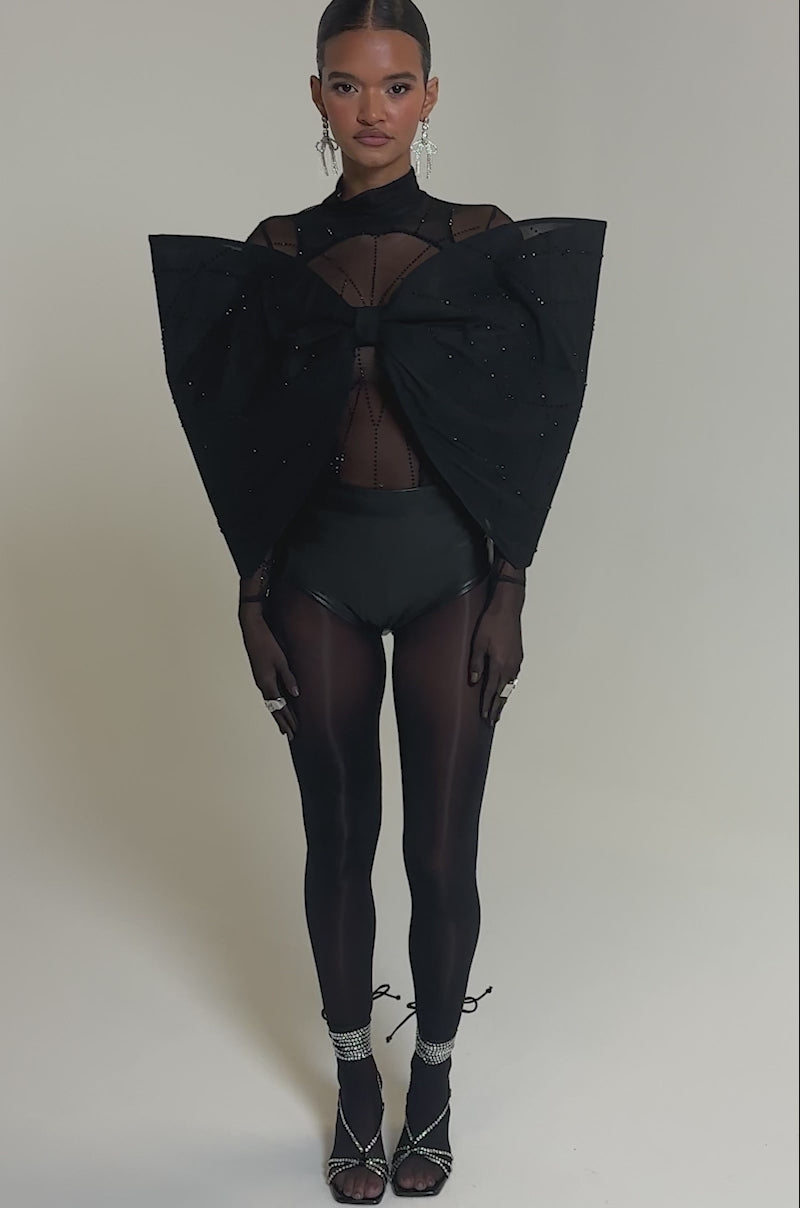 Video of woman modeling the SPIDER GIRL BOW TOP, a sheer black top with a sculptural studded bow, styled with black high-waisted bottoms, sheer gloves, bold rings, and long silver earrings against a neutral background.