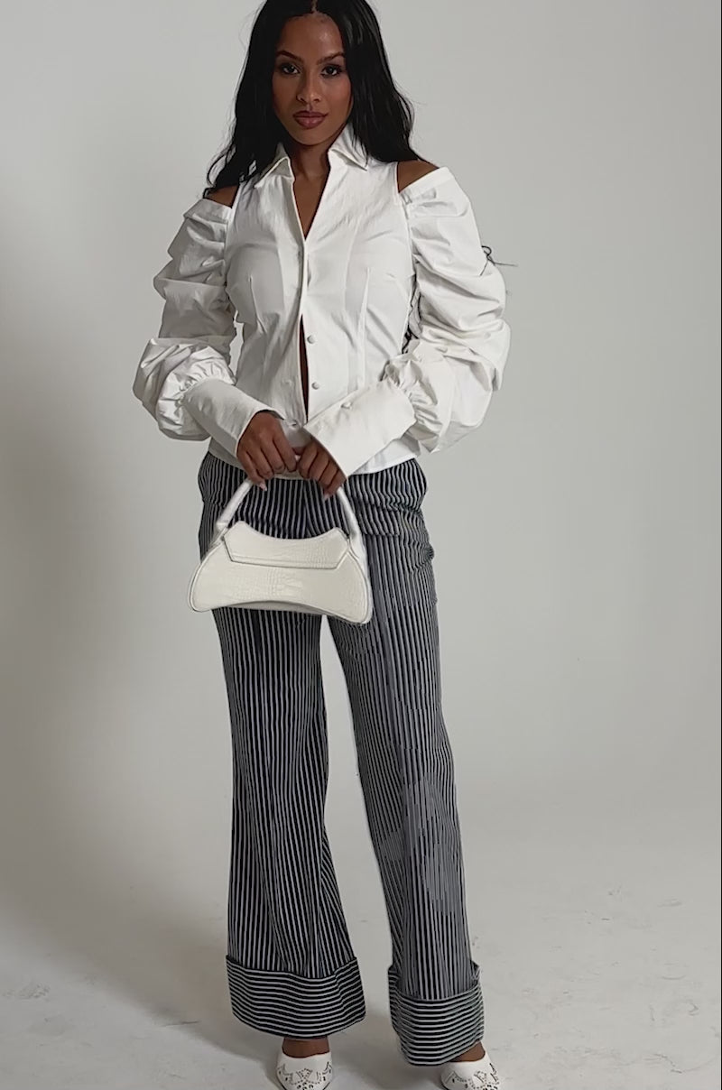 Video of woman modeling the CRAZY IN LOVE WHITE BUTTON DOWN BLOUSE with long, ruched sleeves and pinstriped gray pants.