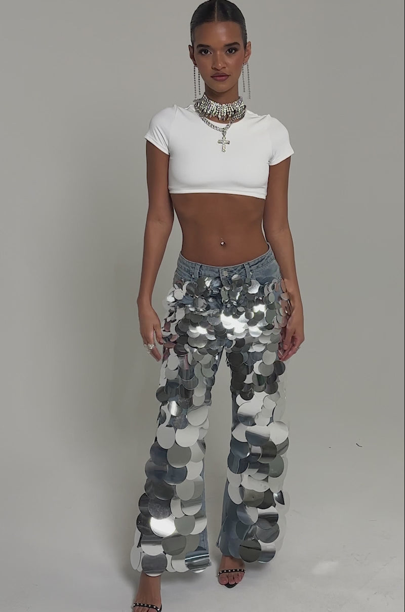 Video of woman modeling the CHECK IT OUT SEQUIN DENIM PANT—vintage-wash blue jeans with large silver sequins—paired with a white crop top, layered necklaces, black heels, and a sleek updo, posing against a plain background.