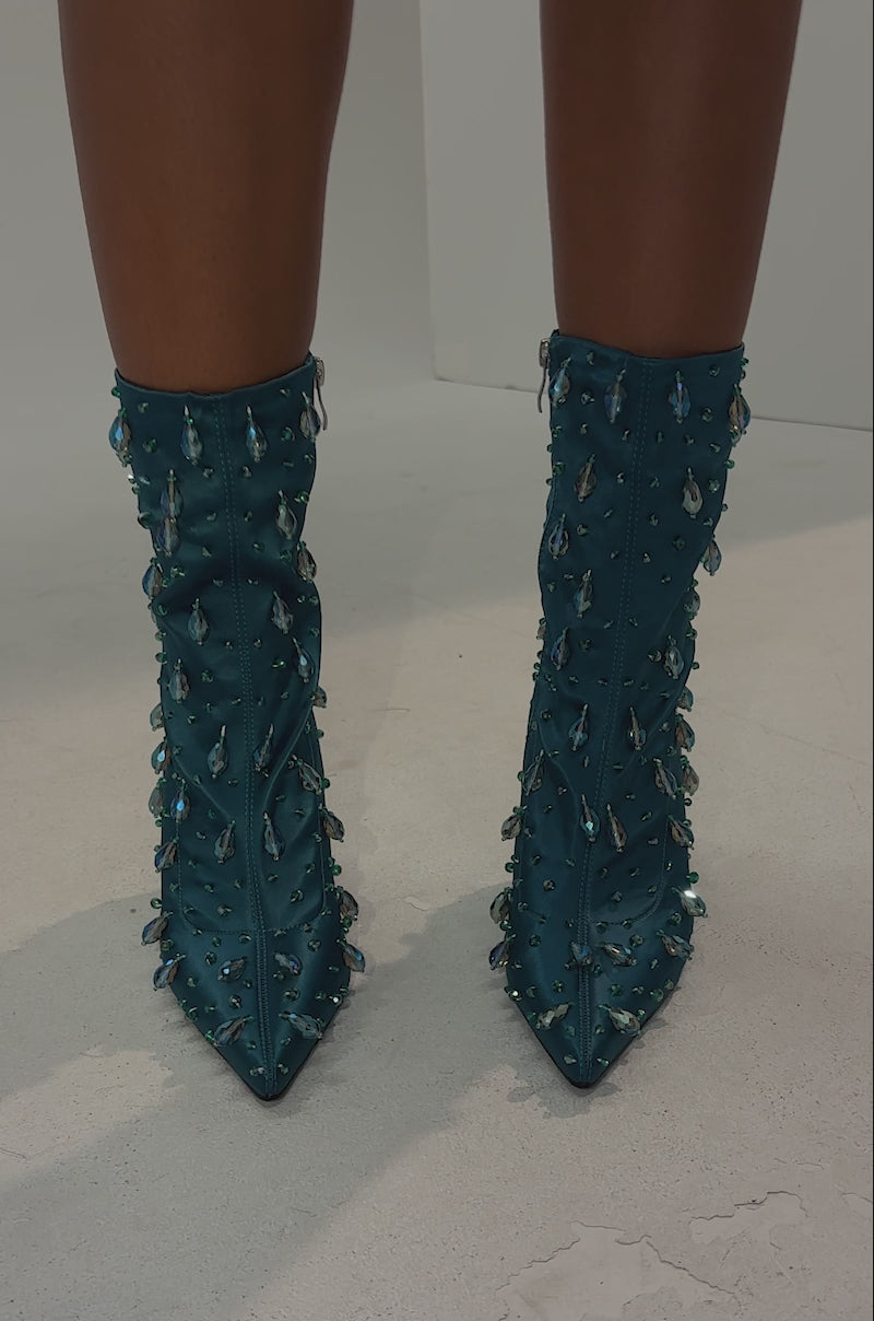 A video of a person in navy shorts showcaseing the AZALEA WANG IZIDOR TEAL EMBELLISHED RHINESTONE BOOTIE against a dark background, highlighting their legs and the sparkling, rhinestone-studded footwear.