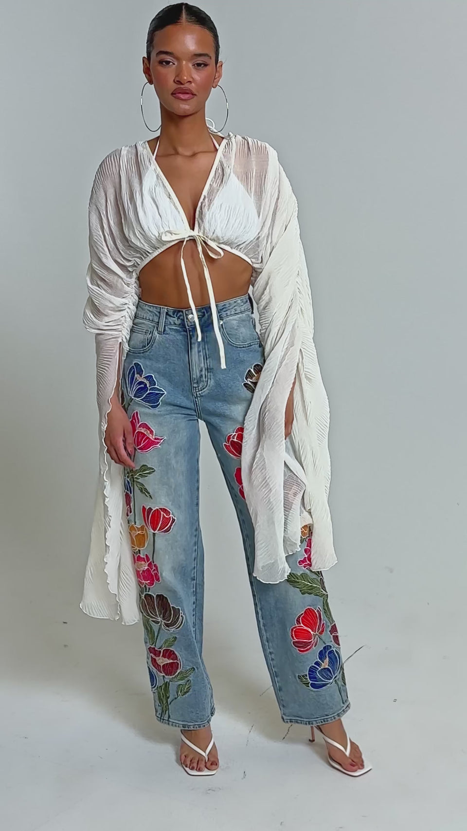 Video of woman modeling the FLY AWAY CHIFFON BLOUSE—a sheer, pleated top with wide sleeves—styled with a white halter bralette, embroidered high-waisted jeans, heeled sandals, and her hair pulled back.