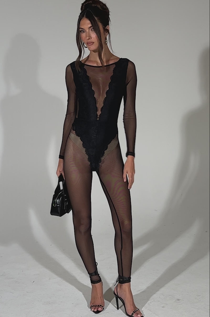 A video of a woman standing against a plain background wearing the ALL ABOUT ME MESH JUMPSUIT—a sheer black one-piece with a deep V neckline—styled with a small black handbag and high-heeled sandals.