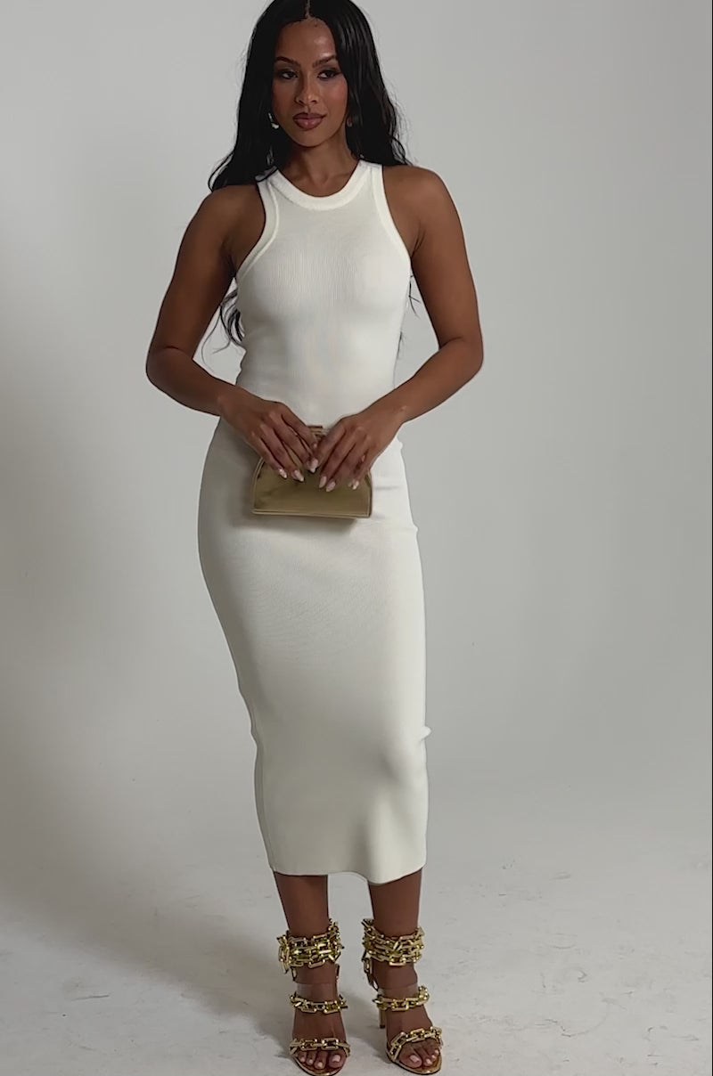 Video of woman modeling the CUTIE PIE KNIT MIDI DRESS IN WHITE holding a small gold clutch and wearing gold chain-detailed high-heeled sandals. She poses against a plain light background, her long wavy dark hair flowing freely.