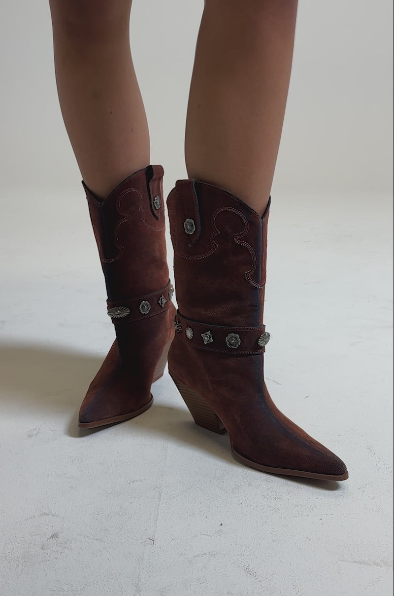 A video of a person wearing the AZALEA WANG NEITH BURNT ORANGE DENIM WESTERN BOOTIE, featuring mid-calf height, pointed toe, decorative silver conchos, and a slanted heel, stands on a neutral background.