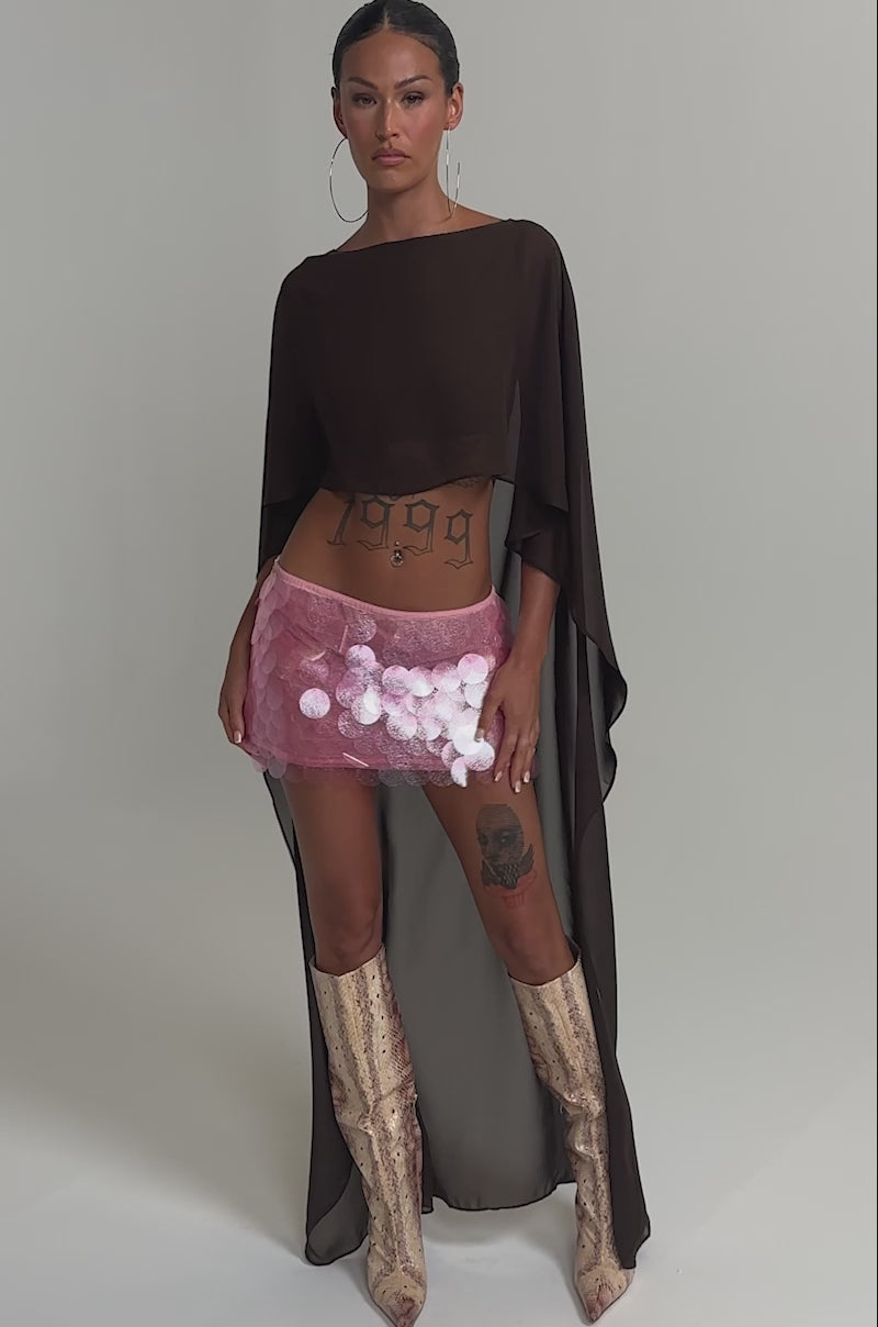 Video of woman modeling the IT GIRL BLOUSE IN BROWN, a sheer chiffon top with a high-low hem, styled with a pink sequin mini skirt, snakeskin knee-high boots, hoop earrings, a wide belt, and her midriff tattoo visible against a plain background.