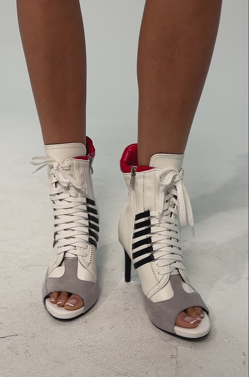 A video of a model wearing the AZALEA WANG EOWYN WHITE MULTI OPEN TOE STILETTO BOOTIE. The bootie is a high-heeled, lace-up ankle boot featuring white leather with black stripes, a red top accent, grey suede toe, and an open toe design.