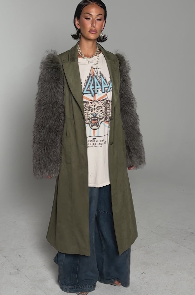 A video of a woman modelling the LOLITA FUR SLEEVE TRENCH in olive green with faux fur sleeves, paired with a white graphic tee and wide-leg blue jeans, standing barefoot against a plain light gray background.