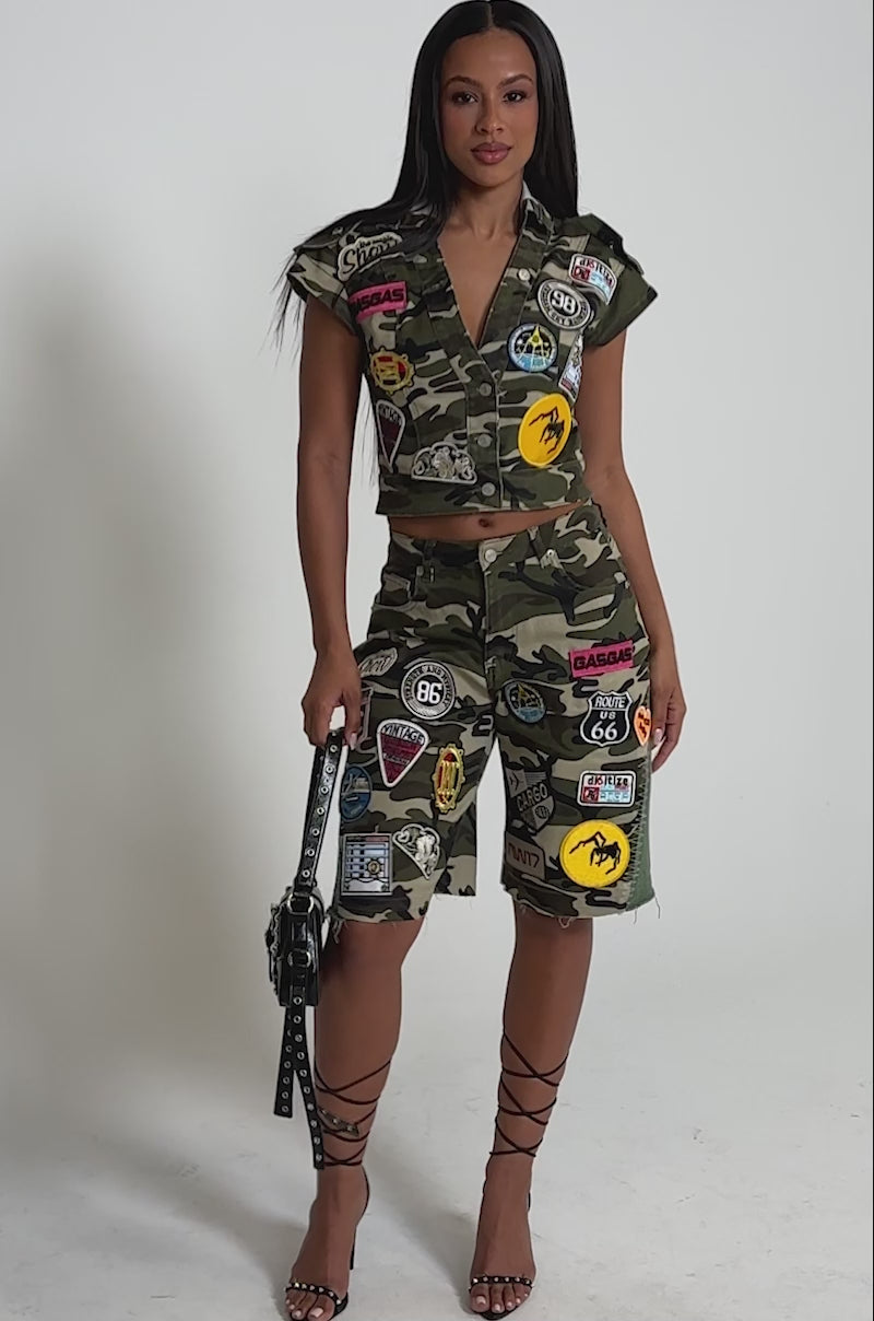 A video of a woman in the DOWN FOR SOME FUN CAMO BUTTON DOWN, styled with camo shorts, colorful patches, a studded handbag, and high-heeled lace-up sandals.