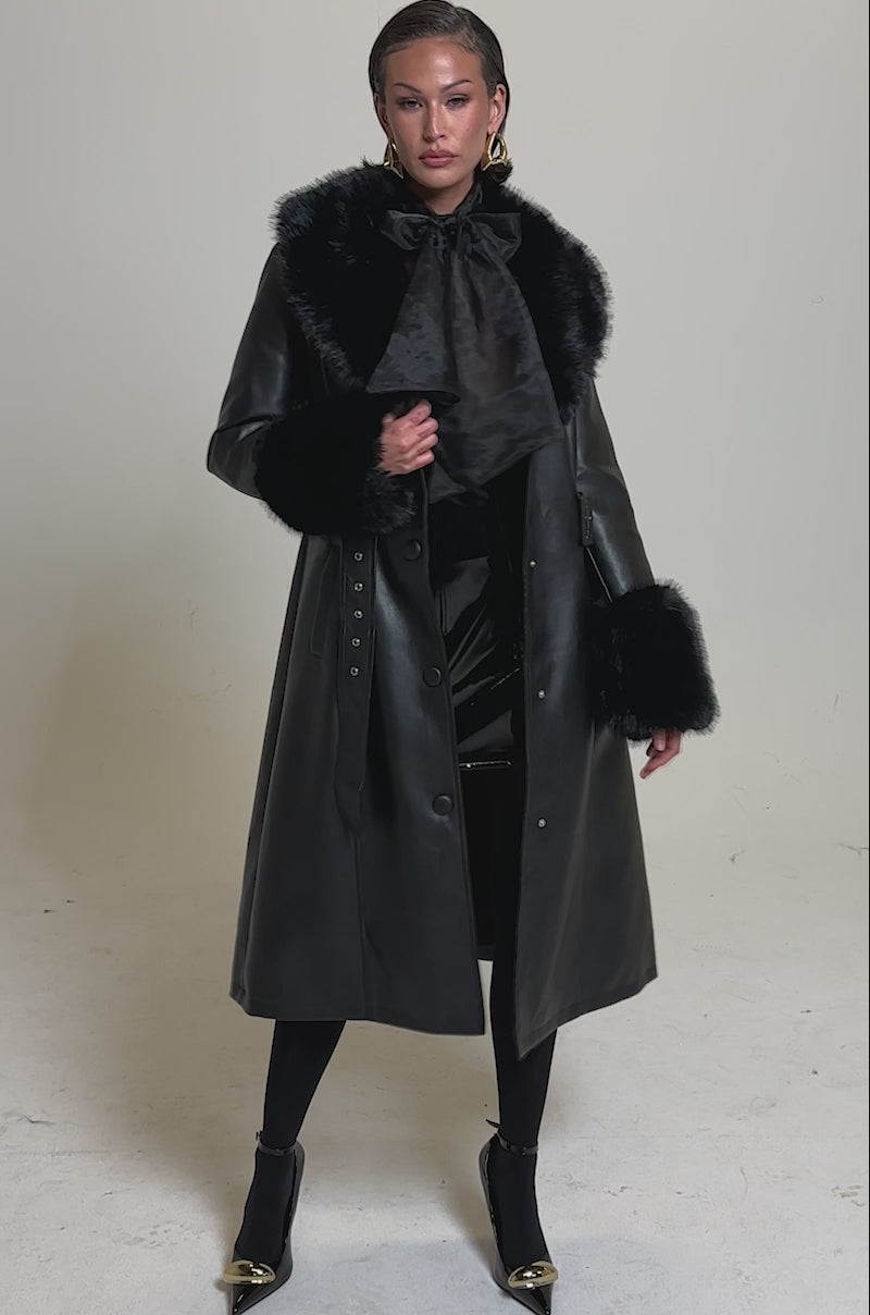 A video of a woman in the KAYA FUR TRIM TRENCH, a black pleather coat with a wide belt and faux fur on the collar and cuffs, styled with black tights and pointed gold-tip heels against a plain light background.