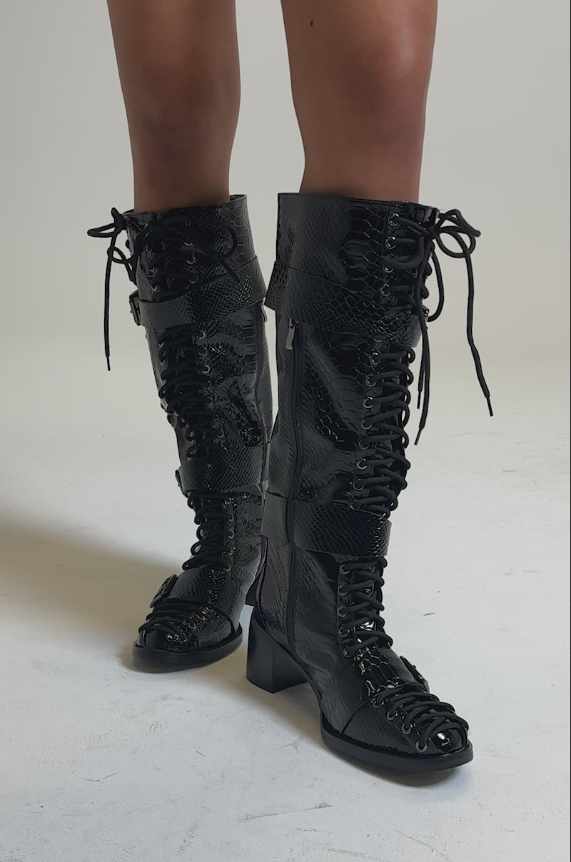 A video of a person wearing the AZALEA WANG MAKAR BLACK CROC LACE UP DETAIL BOOT—knee-high, with buckles and a chunky heel—standing against a neutral background.
