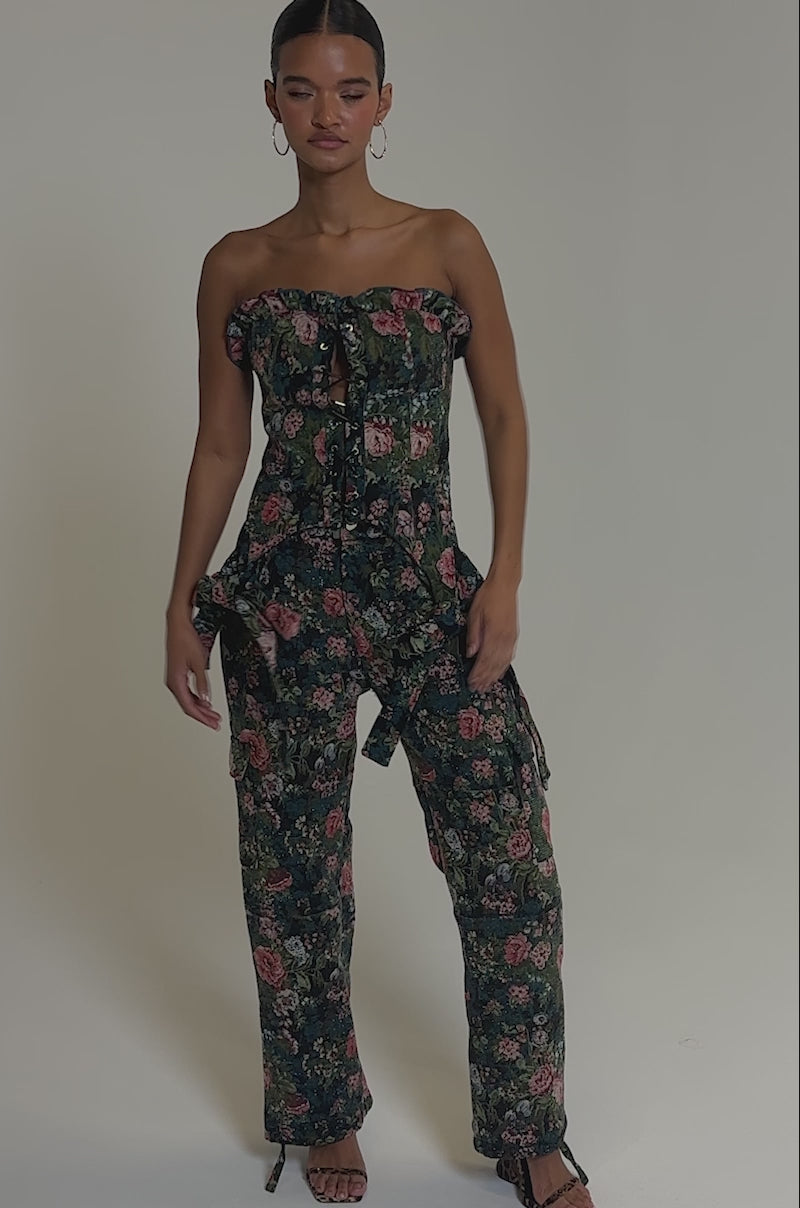 Video of woman modeling the I WANT YOU FLORAL RHINESTONE CORSET, a strapless floral jumpsuit with a lace-up front and tied bows at the hips. She accessorizes with hoop earrings and has her hair pulled back, posing against a plain background.