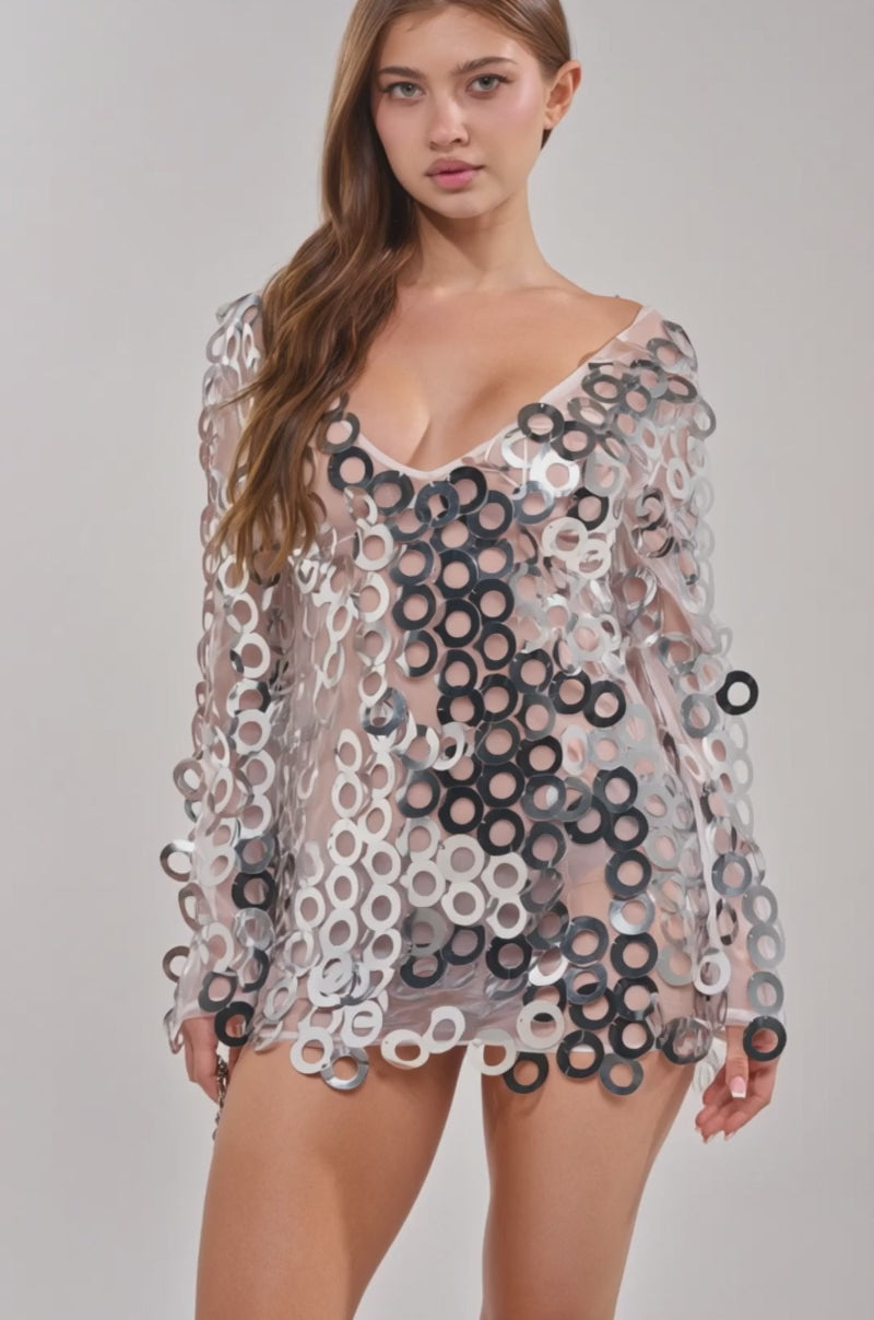 Video of woman modeling the SEE ME SPARKLE SEQUIN MINI DRESS—a sheer, long-sleeved mini dress with bold silver sequins—a woman with long brown hair poses against a light gray backdrop, chicly accessorizing with a small silver handbag.