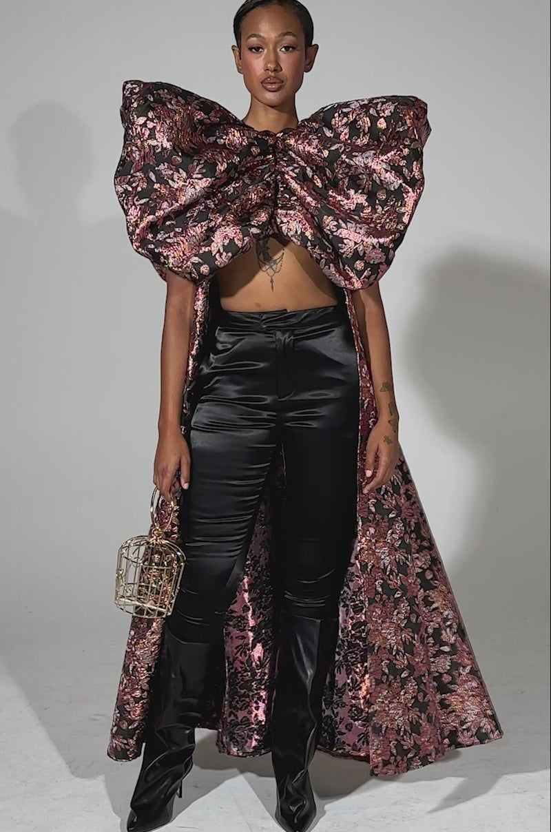 A video of a woman wearing the DIVINE INTERVENTION BROCADE TOP—featuring dramatic pink and black florals, puffed shoulders, and a large matching bow—paired with black pants, heels, and a birdcage-shaped purse against a light gray background.