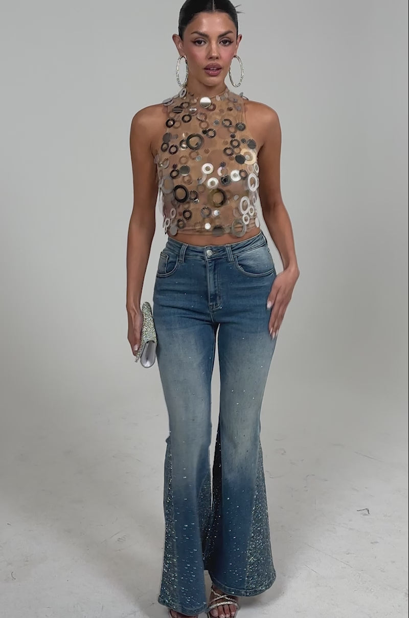 Video of woman modeling the BLING BLING FLARE DENIM, a sleeveless sheer halter top with circular accents, silver heels, large hoop earrings, and a small silver clutch.