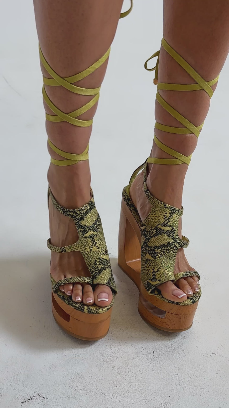 Video of woman modeling the AKIRA GOLD RIN GREENSNAKE NOVELTY LACE UP WEDGE SANDAL, featuring yellow snakeskin straps that wrap around the calves and chunky wooden heels, styled with denim shorts against a plain light background.