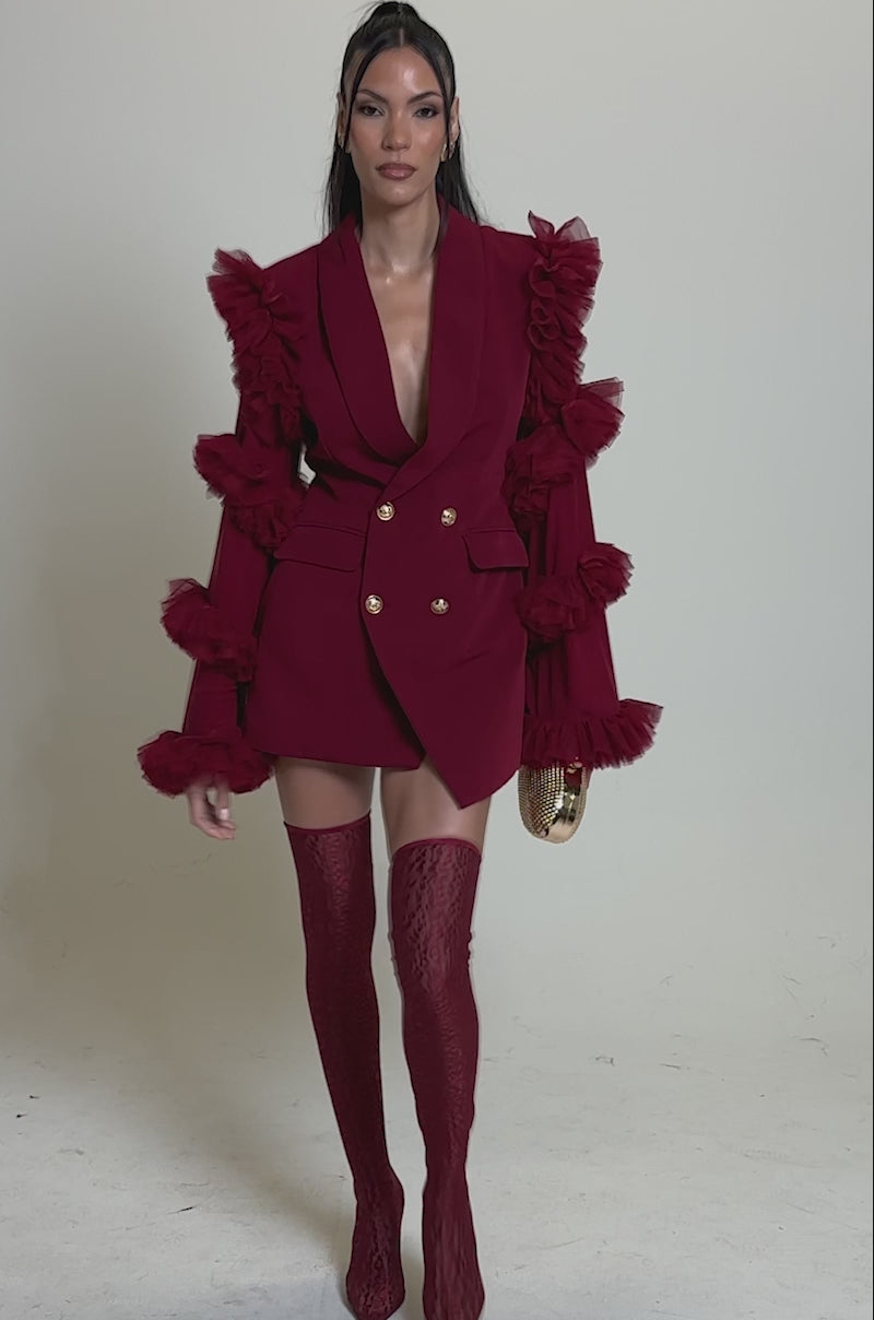 Video of woman modeling the LIVING OVER THE TOP RUFFLE SLEEVE BLAZER MINI DRESS IN BURGUNDY, styled with ruffled tulle sleeves, matching thigh-high stockings, gold earrings, a round clutch, and long straight hair.