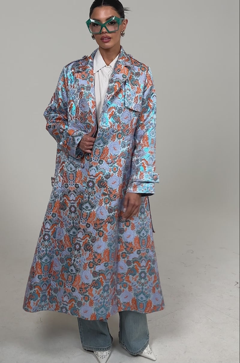 Video of woman modeling the ST. GERMAIN BROCADE TRENCH, turquoise glasses, a white shirt, blue jeans, and white heels.