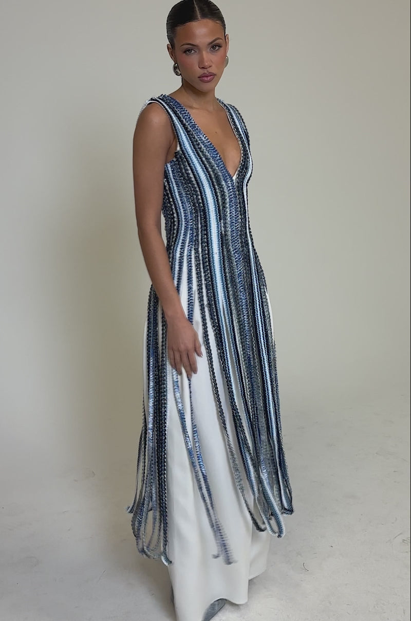 Video of woman modeling the TIED DOWN BRAIDED ROPE TOP—a sleeveless white top with long vertical blue and white fringe and a deep V-neckline—styled with silver heels against a plain light background.