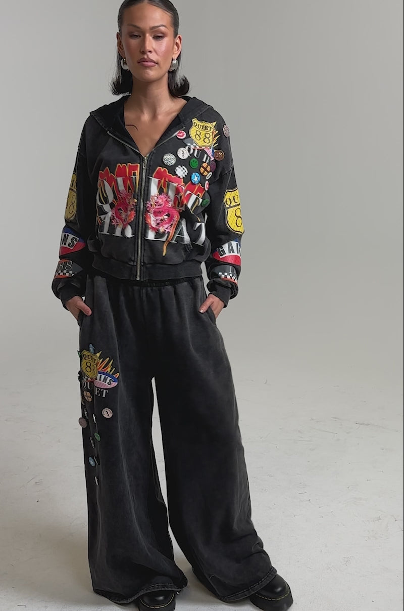 A video of a woman in the ANOTHER ONE WIDE LEG JOGGER and matching zip-up hoodie, both featuring colorful graphic patches. 