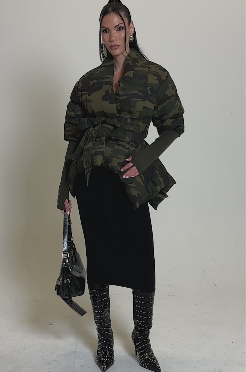 Video of woman modeling the RIB SLEEVE PEPLUM PUFFER IN CAMO, an oversized belted jacket with wide, ribbed sleeves, styled over a black buttoned skirt. Her long dark hair is pulled back and she accessorizes with large earrings and a black strap in hand.