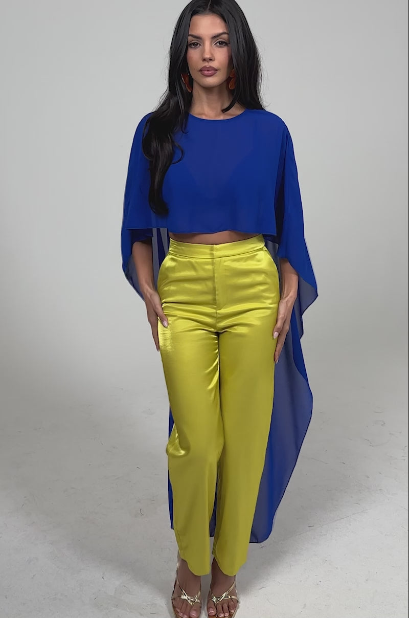 Video of woman modeling the COOL GIRL CHIFFON BLOUSE IN ROYAL BLUE styled as a cape over a crop top and yellow pants, paired with gold sandals.