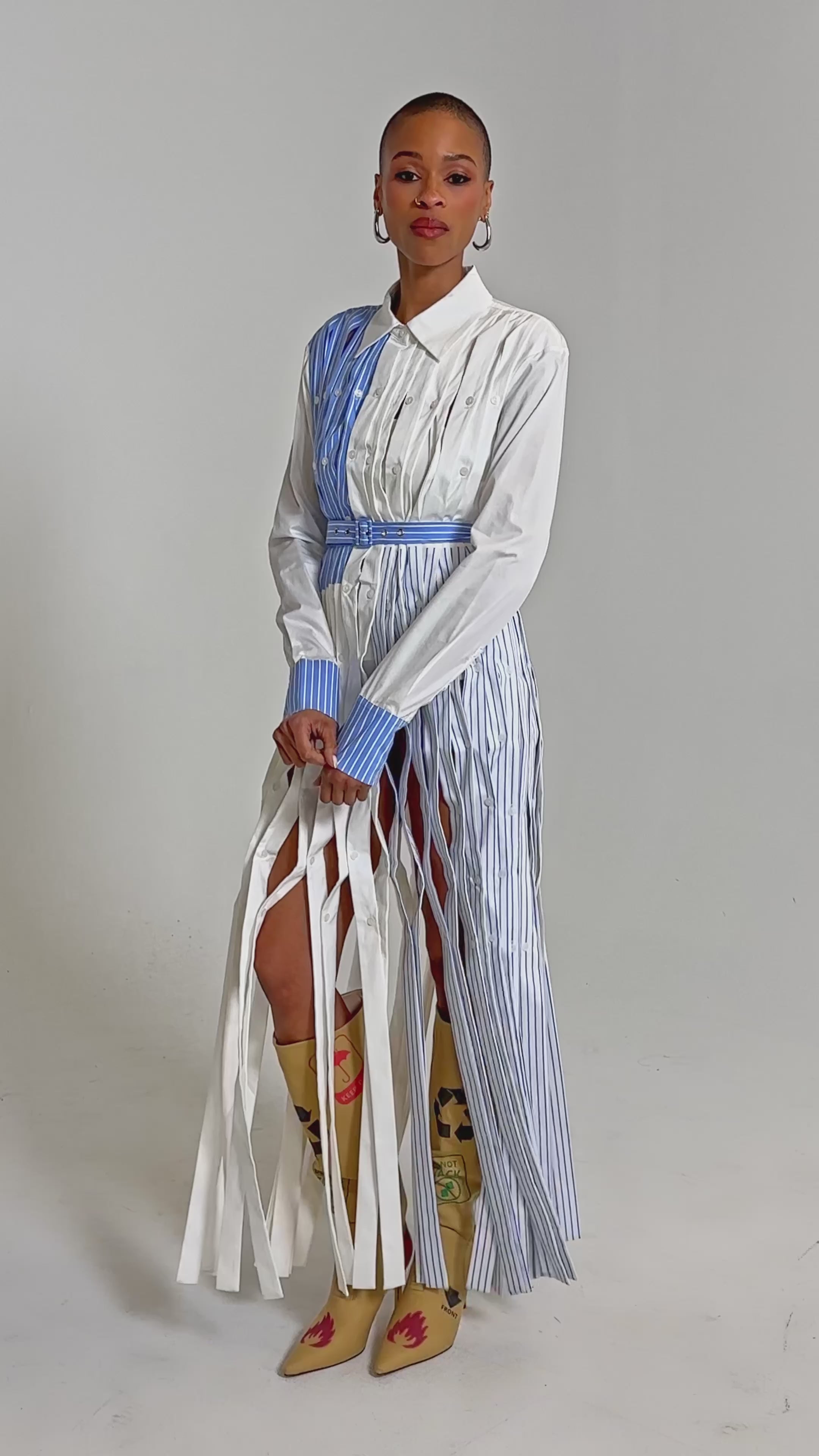Video of woman modeling the NOT MY MANS BUTTON UP SHIRT MAXI DRESS with vertical blue stripes and fringe accents, styled with tall tan floral boots, statement earrings, and a confident look against a plain background.