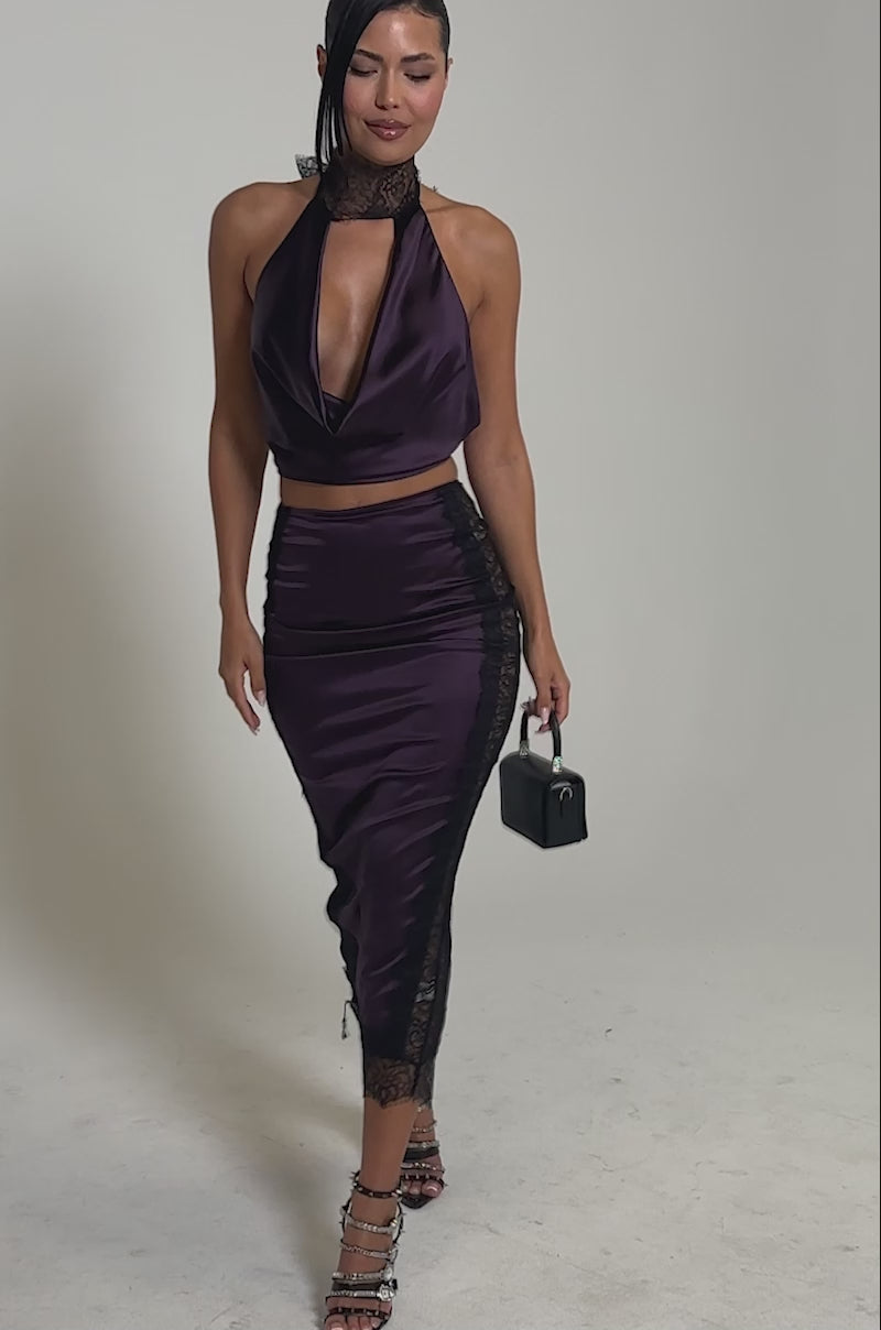Video of woman modeling the GOLDEN GIRL SATIN MAXI SKIRT, a plum satin piece with lace trim. She pairs it with a shiny purple halter top, strappy silver heels, and a small black handbag. Her hair is styled to one side against a plain white background.