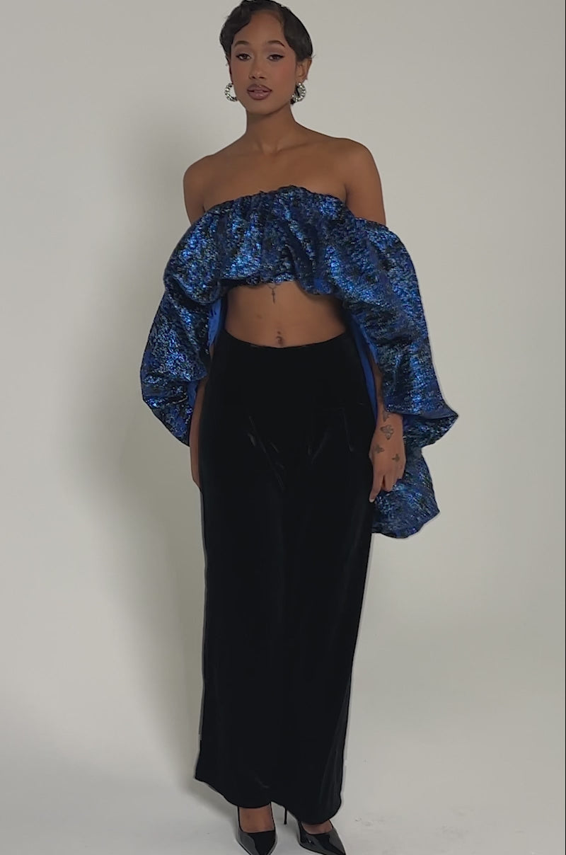 Video of woman modeling the MELANI OFF THE SHOULDER HI-LOW BROCADE BLOUSE, paired with black velvet pants and silver rings and earrings against a neutral background.