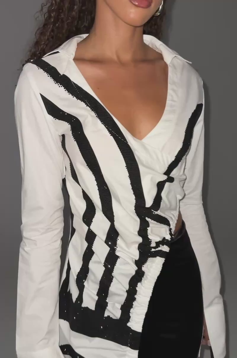 Video of woman modeling the GET MY CLOSE UP EMBELLISHED BUTTON DOWN SHIRT—a long white top with bold black abstract face art—paired with black shiny fitted pants, sandals, and a small black handbag.
