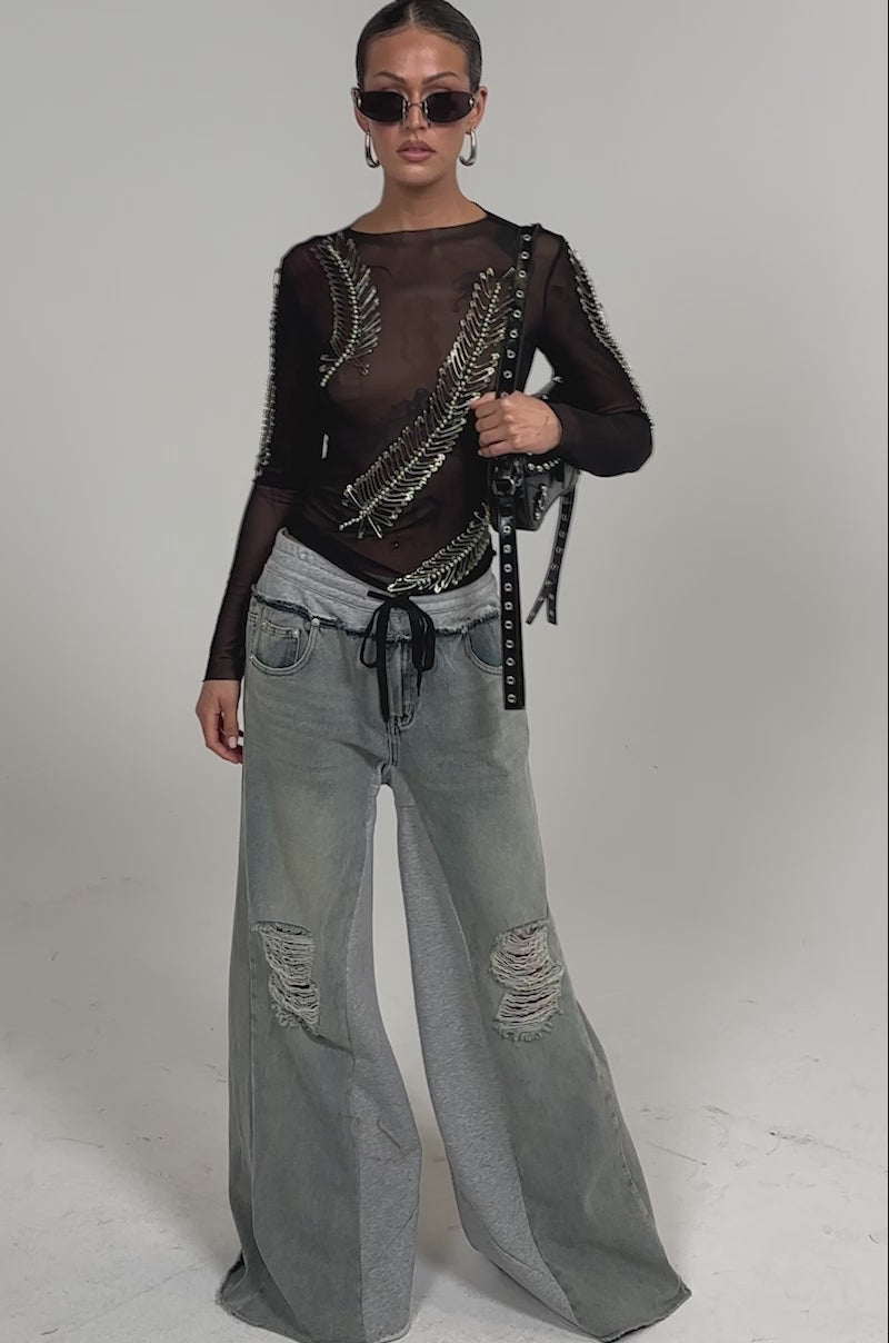 Video of woman modeling the PINNED DOWN MESH TOP with safety pin accents, gray sweatpants layered over blue jeans, hoop earrings, and sunglasses—an edgy look ideal for nightlife fashion.