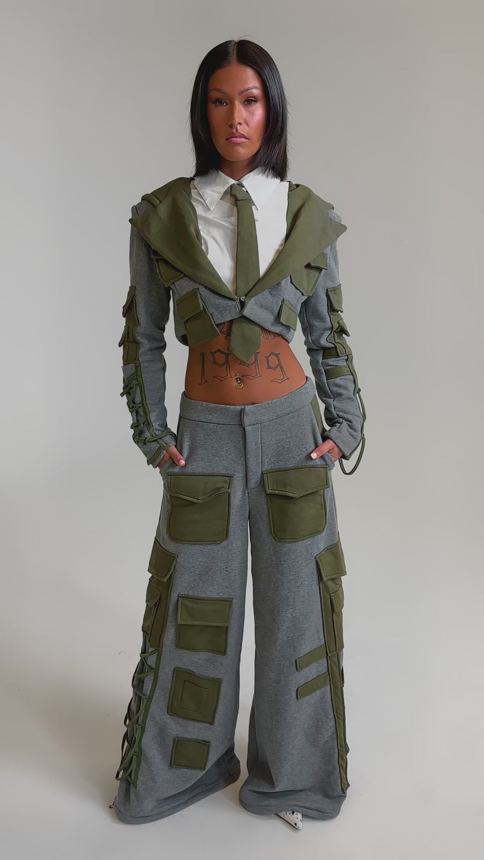 A video of a woman in the CRAZY IN LOVE BLOUSE, paired with a cropped grey jacket with olive green accents and matching wide-leg utility pants, revealing her midriff for an edgy streetwear look.