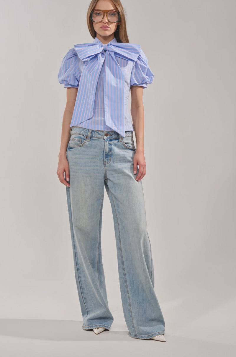 Video of woman modeling the BOW DOWN SHORT SLEEVE BUTTON DOWN—light blue striped with puffed sleeves and a big bow tie—paired with light blue jeans and large, amber-tinted glasses against a plain background.
