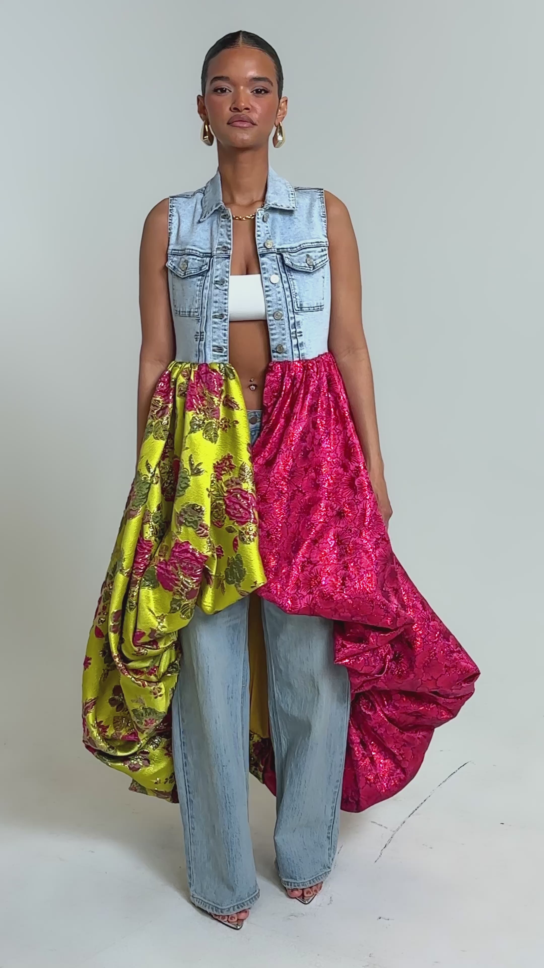 Video of woman modeling the ON THE HIGH LOW DENIM VEST, with a yellow floral brocade, pink ruffled skirt over blue jeans, large hoop earrings, and open-toe shoes—perfect for bold statement occasion 