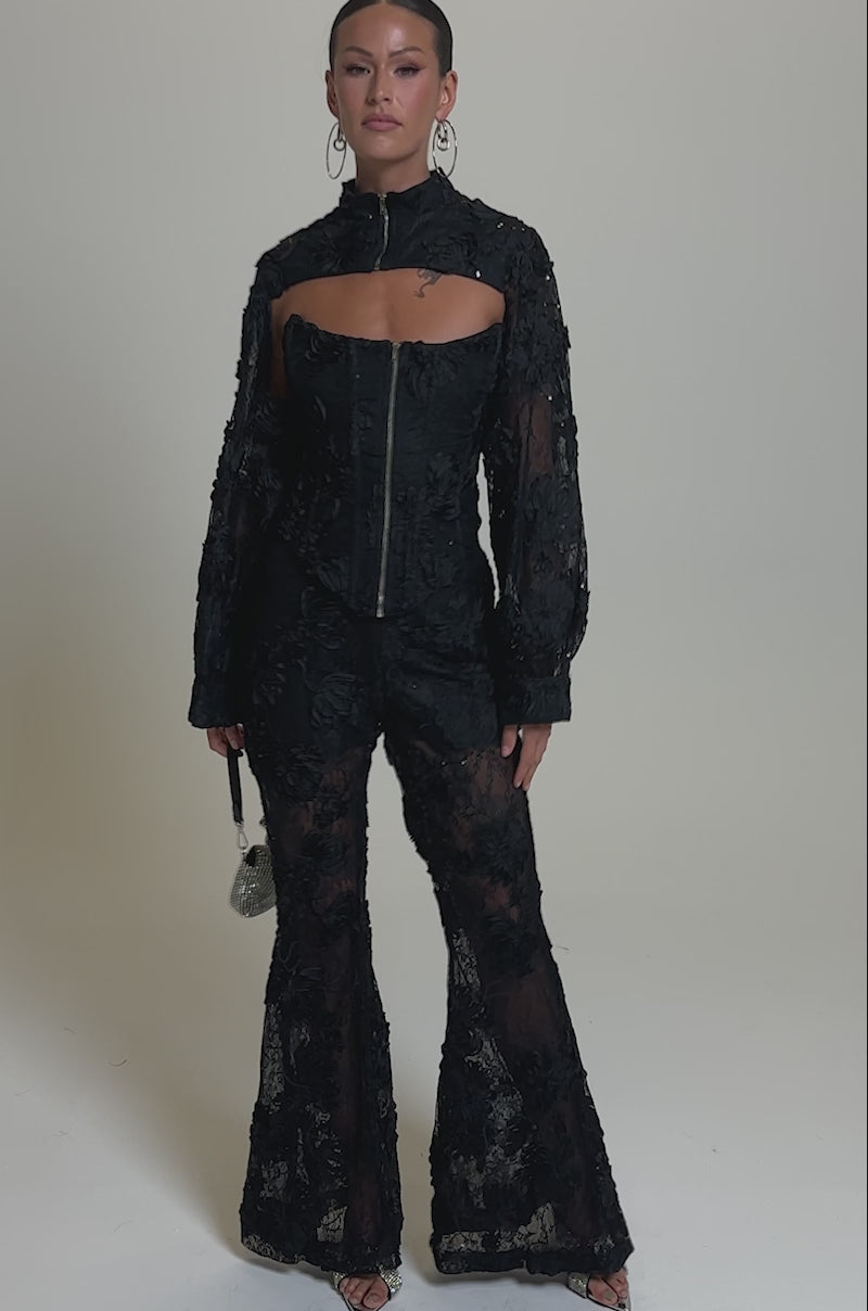 A video of a woman in the LET IT GO LACE CORSET, pairing it with flared pants. She holds a small silver purse and accessorizes with hoop earrings, heels, and slicked-back hair.