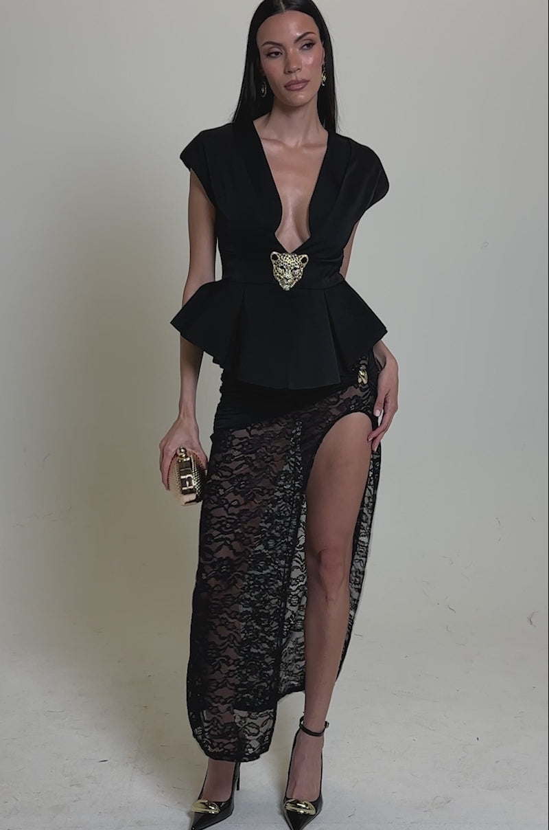 A video of a woman wearing the FIERCE BLOUSE with a deep V-neck and peplum hem, styled with a gold brooch, lace high-slit skirt, black heels with gold tips, and a small gold clutch.