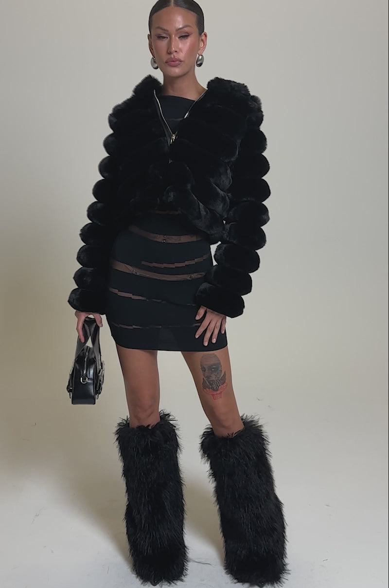 A video of a woman wearing the POPCORN FAUX FUR IN BLACK, paired with a sheer black mini skirt with cutouts, black fuzzy knee-high boots, and a small black handbag.