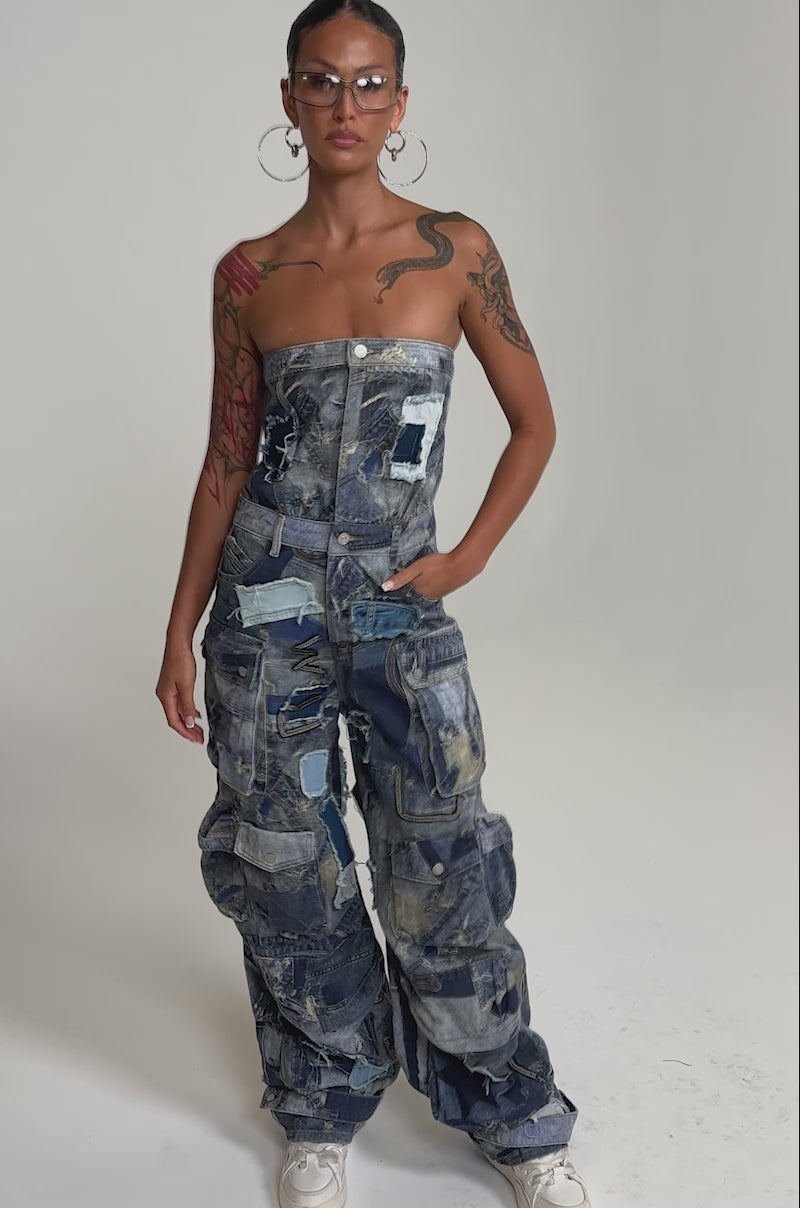 A video of a model wearing the PATCHWORK DENIM ON DENIM JUMPSUIT with wide legs and large pockets in mixed denim shades, paired with white sneakers, hoop earrings, and clear glasses.