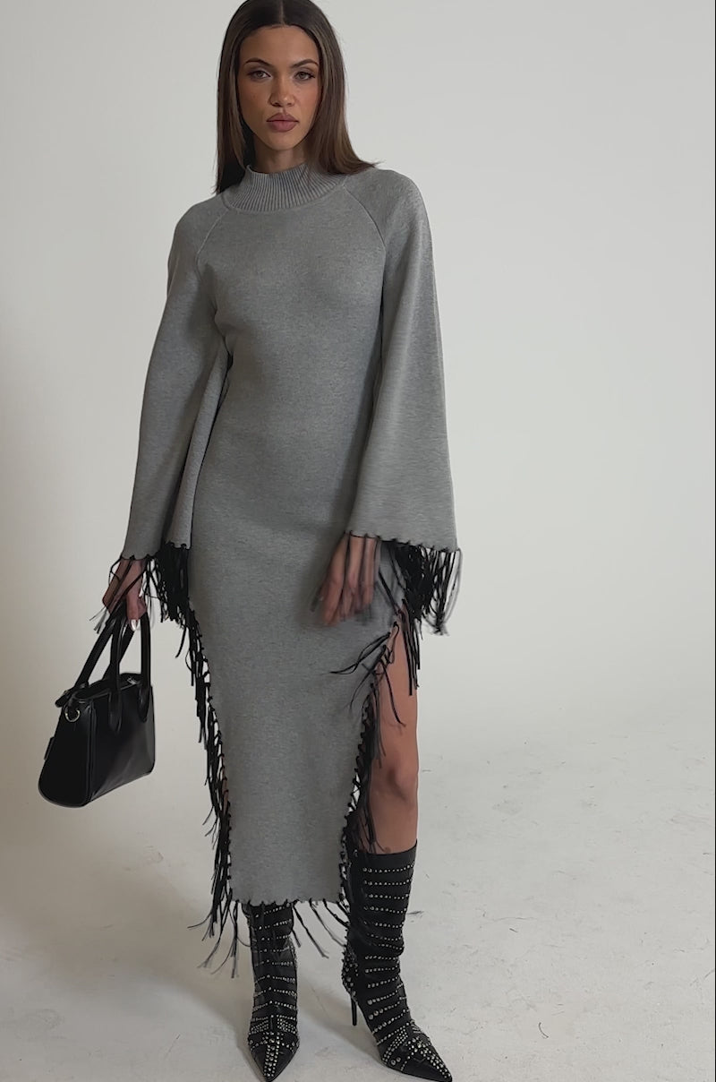 Video of woman modeling the SWEATER WEATHER KNIT MIDI DRESS IN GREY, featuring subtle faux leather fringe, styled with studded black boots and a black handbag. The plain white background showcases her chic, modern look.
