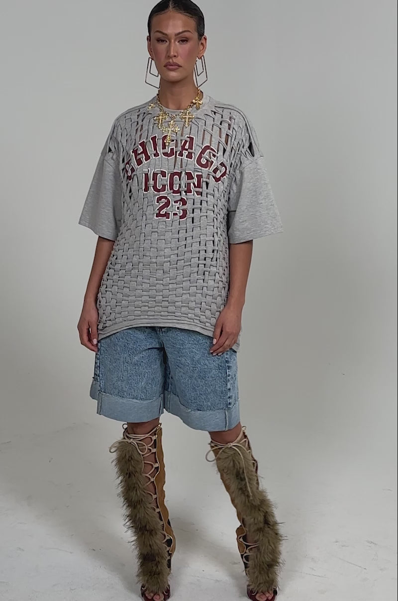 Video of woman modeling the DISTRESSED FOR LIFE OVERSIZED TEE, a grey graphic shirt with Chicago lettering and "ICON 23" print, featuring a cutout overlay design. She pairs it with large hoop earrings and blue denim shorts against a plain background.