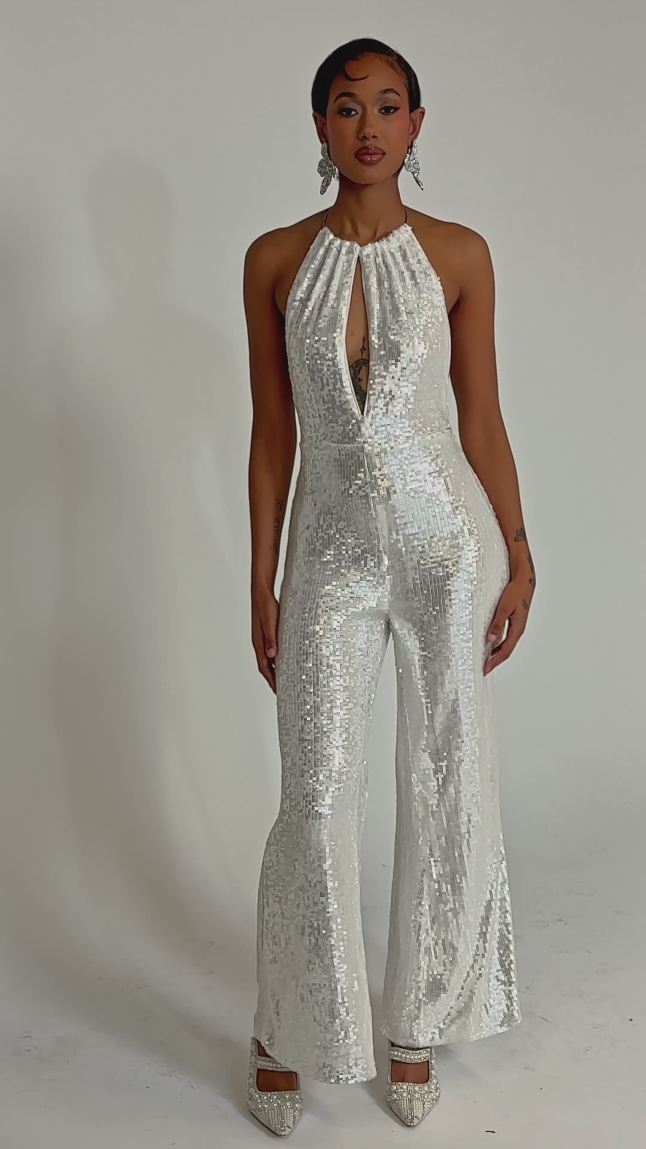 A video of a woman in the ICE QUEEN SEQUIN JUMPSUIT, a sleeveless white sequin piece with a halter neckline and flared legs. She completes the look with short hair, statement earrings, and pointed-toe heels.