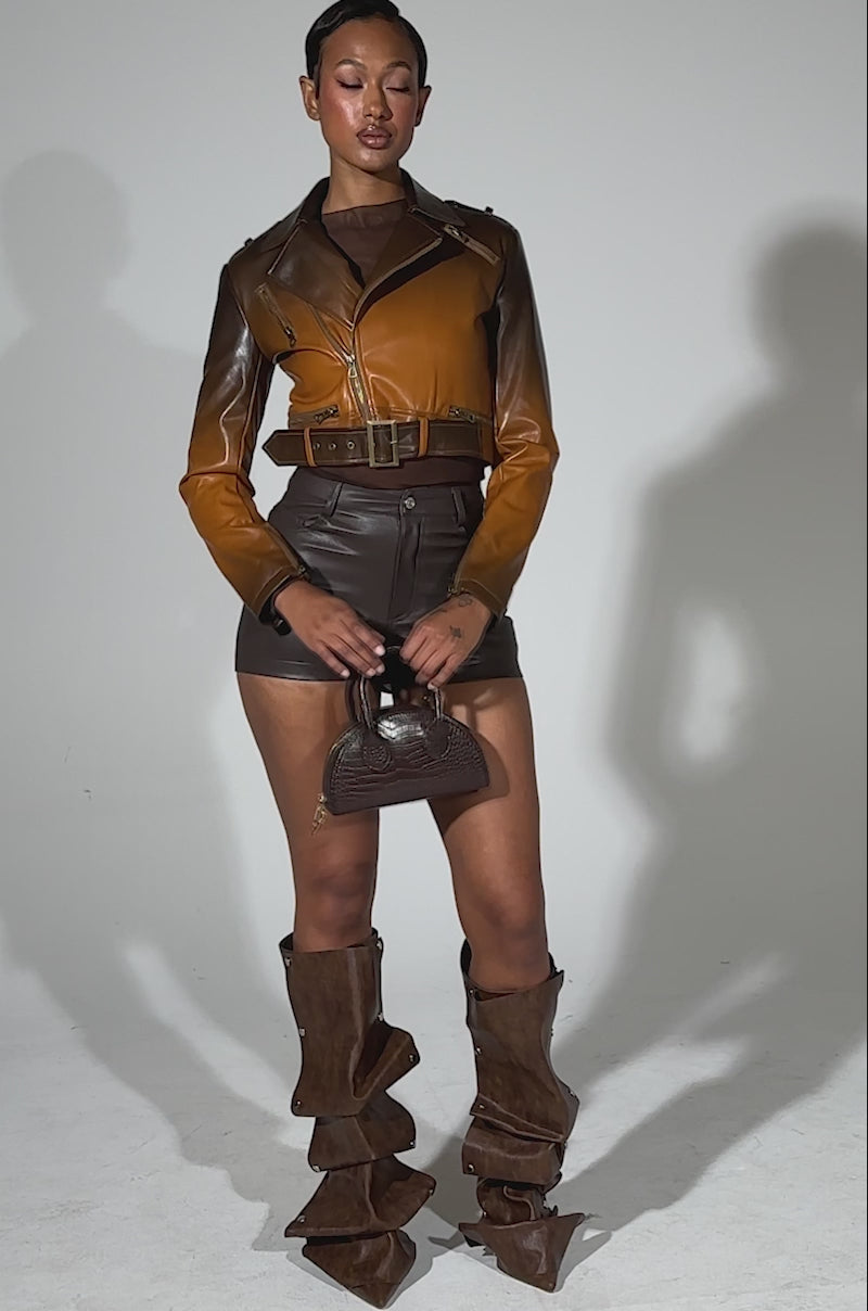 A video of a person wearing high-rise brown faux leather shorts (CALLIE FAUX LEATHER SHORT IN BROWN), a brown leather jacket, slouchy brown knee-high pointed-toe boots, and holding a small brown handbag. 