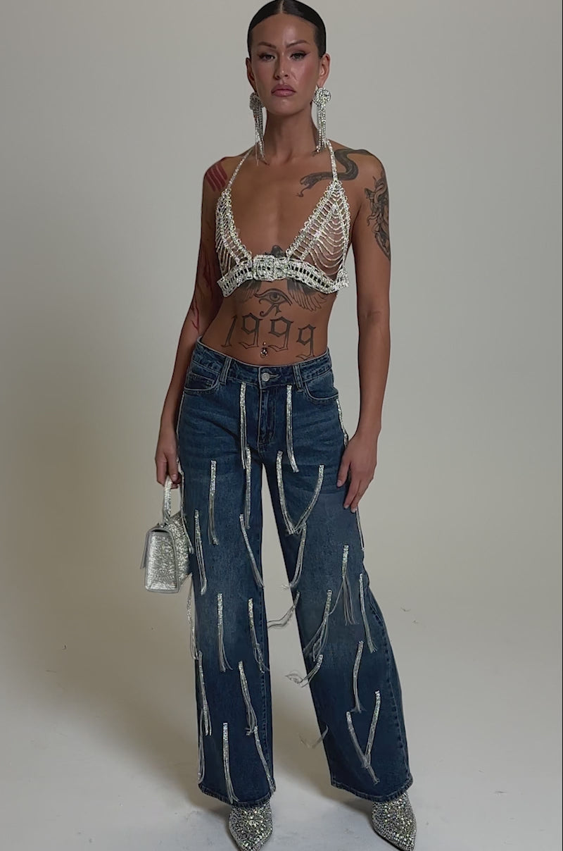 A video of a person in the DRIPPED OUT RELAXED DENIM with rhinestone embellishments, a pearl and chain bralette, glittery heels, large dangling earrings, silver handbag, and visible arm and torso tattoos.